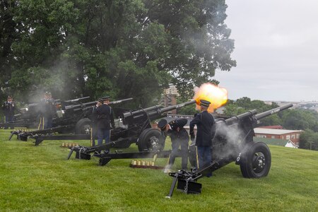 Army soldiers in dark ceremonial uniforms are firing black cannons that are placed on a green lawn.