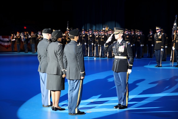 Three soldiers in dark service dress uniforms are facing another soldier who is saluting them. They are standing on a blue colored floor and in the background are soldiers in ceremonial uniforms standing in rows (formation).