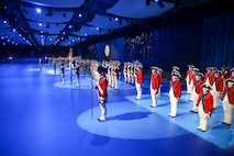 Soldiers in various uniforms, including Revolutionary War-era red with blue trim and white pants, are standing in row (formations) along a wall in large room with spotlights shining on them.