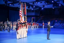 A soldier in a dark ceremonial uniform is pointing a sword out in front of him. He is standing in front of a color guard that is dressed in Revolutionary War-era uniforms while carrying the US flag and the Army flag.