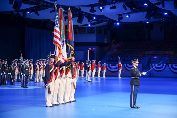 A soldier in a dark ceremonial uniform is pointing a sword out in front of him. He is standing in front of a color guard that is dressed in Revolutionary War-era uniforms while carrying the US flag and the Army flag.
