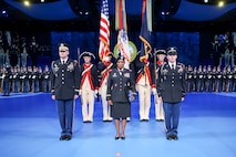Army solders are dressed in dark service uniforms in front of a color guard that is holding the US flag and the Army flag. In the far background are rows of Soldiers standing in formation wearing ceremonial uniforms.