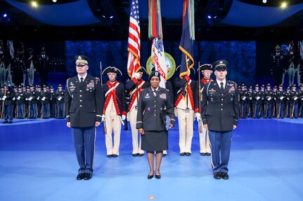 Army solders are dressed in dark service uniforms in front of a color guard that is holding the US flag and the Army flag. In the far background are rows of Soldiers standing in formation wearing ceremonial uniforms.