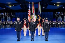 Army solders are dressed in dark service uniforms in front of a color guard that is holding the US flag and the Army flag. In the far background are rows of Soldiers standing in formation wearing ceremonial uniforms.
