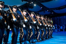 Army Soldiers in dark ceremonial uniforms are marching in rows (formation) with ceremonial rifles on their shoulders.