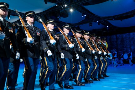Army Soldiers in dark ceremonial uniforms are marching in rows (formation) with ceremonial rifles on their shoulders.