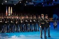 Army soldiers dressed in dark ceremonial uniforms are marching in rows (formation) with ceremonial rifles on their shoulders. One of the Soldiers in the front is holding out a guidon flag in front of him.