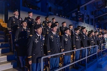 Dozens of Army Soldiers dressed in dark service uniforms are standing at attention and singing from the stadium seats in Conmy Hall.