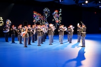 Army Soldiers in dark ceremonial uniforms are performing on brass and percussion instruments. There is a large screen behind them with a projection of their images.