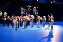Army Soldiers in dark ceremonial uniforms are performing on brass and percussion instruments. There is a large screen behind them with a projection of their images.