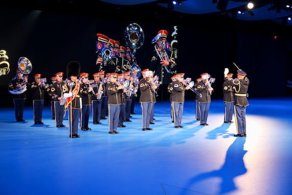 Army Soldiers in dark ceremonial uniforms are performing on brass and percussion instruments. There is a large screen behind them with a projection of their images.