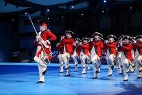 Army soldiers dressed in Revolutionary War-era red with blue trim coats, white pants and tri-cornered hats are marching while playing fifes, bugles and drums. The member in front is holding a long stick.
