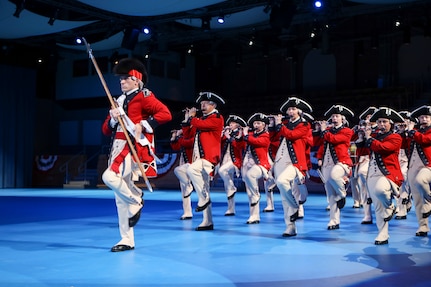 Army soldiers dressed in Revolutionary War-era red with blue trim coats, white pants and tri-cornered hats are marching while playing fifes, bugles and drums. The member in front is holding a long stick.