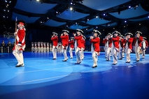 Army soldiers dressed in Revolutionary War-era red with blue trim coats, white pants and tri-cornered hats are marching while playing fifes, bugles and drums. The member in front is holding a long stick.