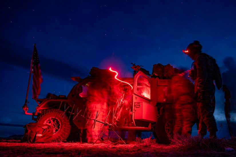 Airmen are seen in a red light’s glow standing next to a military vehicle in a field under a dark blue sky.