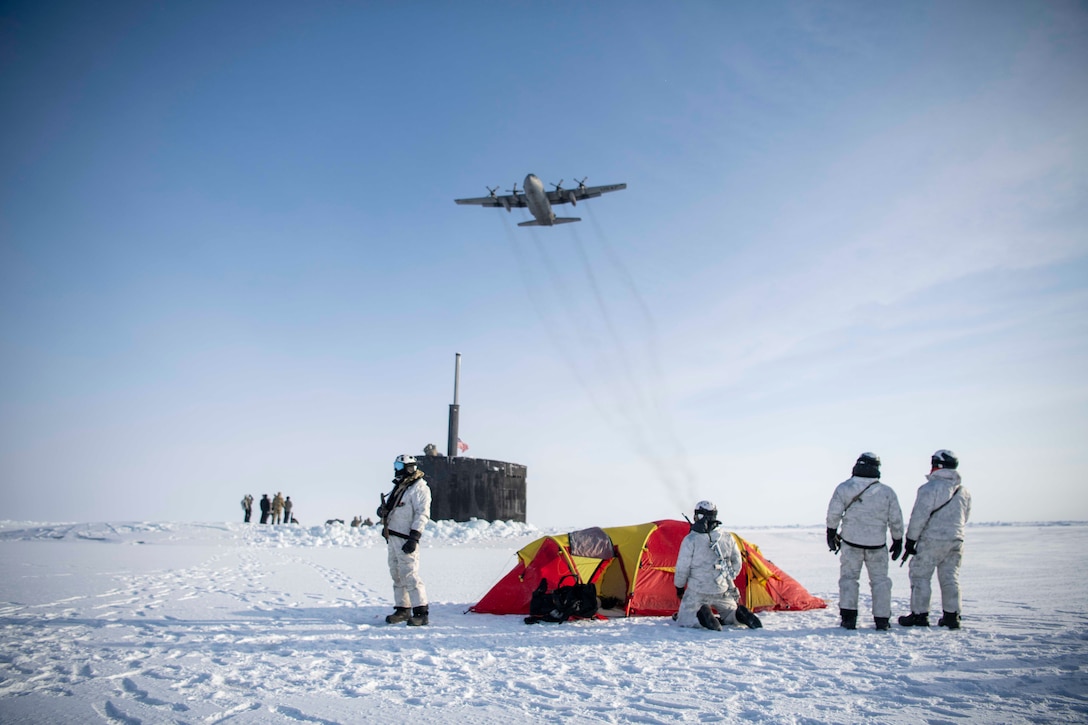A large aircraft flies over a group of service members in winter uniforms standing on the ice next to a tent with the top of a submarine poking through the ice behind them.