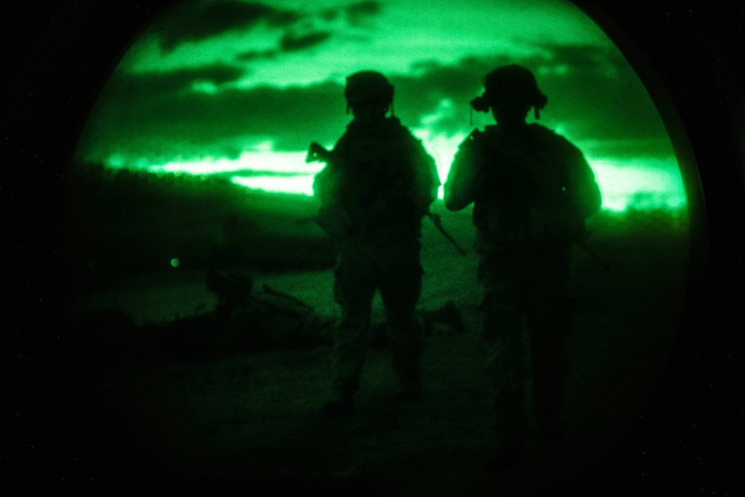 Silhouettes of two soldiers standing together holding weapons outdoors are visible by green light from a night-vision device.