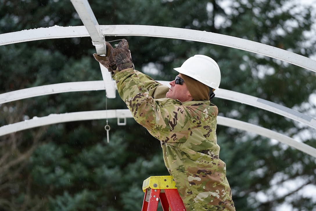 A service member wearing a helmet and standing on a ladder reaches up to install a support beam outdoors.