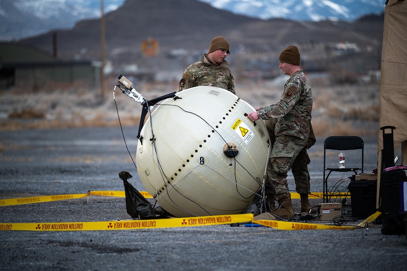 An airman watches as a second one supports a round satellite communications terminal outdoors.