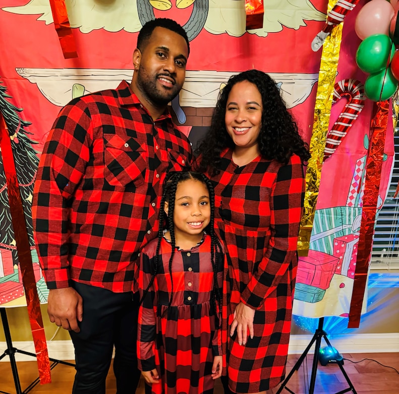 Man, woman, and female child dressed in traditional Christmas colors pose in front of a seasonal backdrop for a holiday family photo