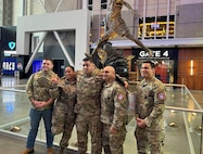 Group of men and women wearing U.S. Army uniforms smile and pose in front of a professional basketball stadium entrance.