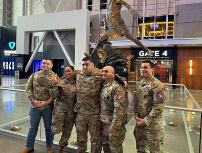 Group of men and women wearing U.S. Army uniforms smile and pose in front of a professional basketball stadium entrance.