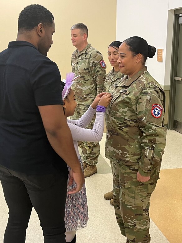 A young girl places a new rank on her Mother's Army uniform during a Military Promotion ceremony while her father and other Army Soldiers stand witness