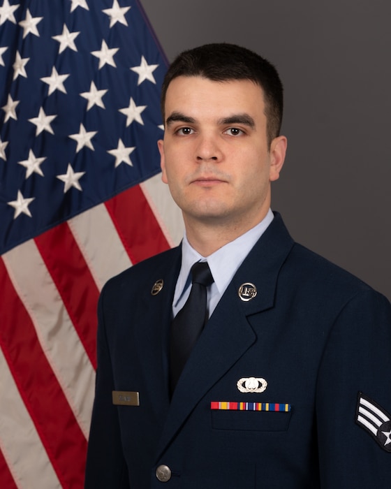 Official photo of SrA Daniel Gelman in front of the American flag. He is wearing his blue service dress uniform.