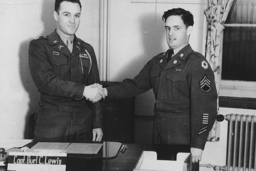 Two service members shake hands while standing behind a desk in this black and white photo.