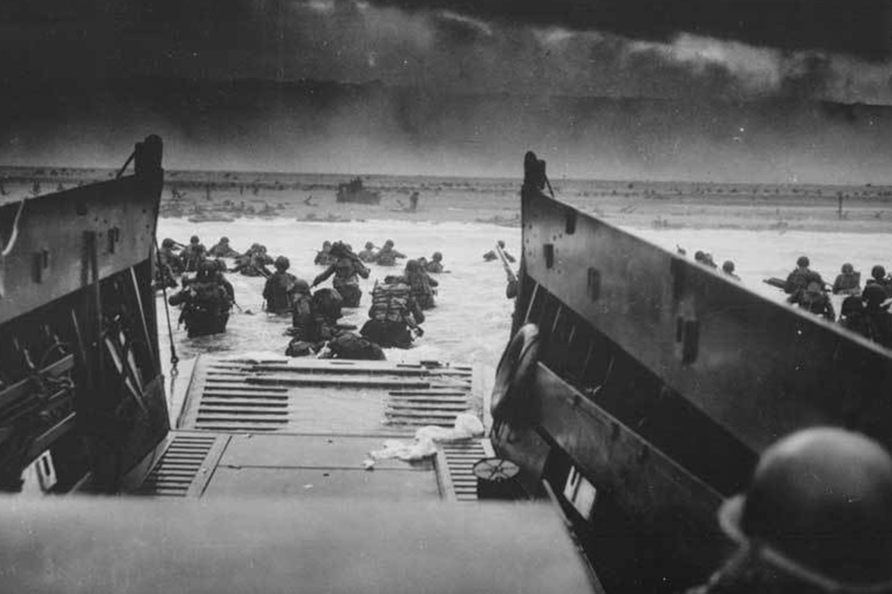 Dozens of service members run into water on a beachhead from a landing craft.