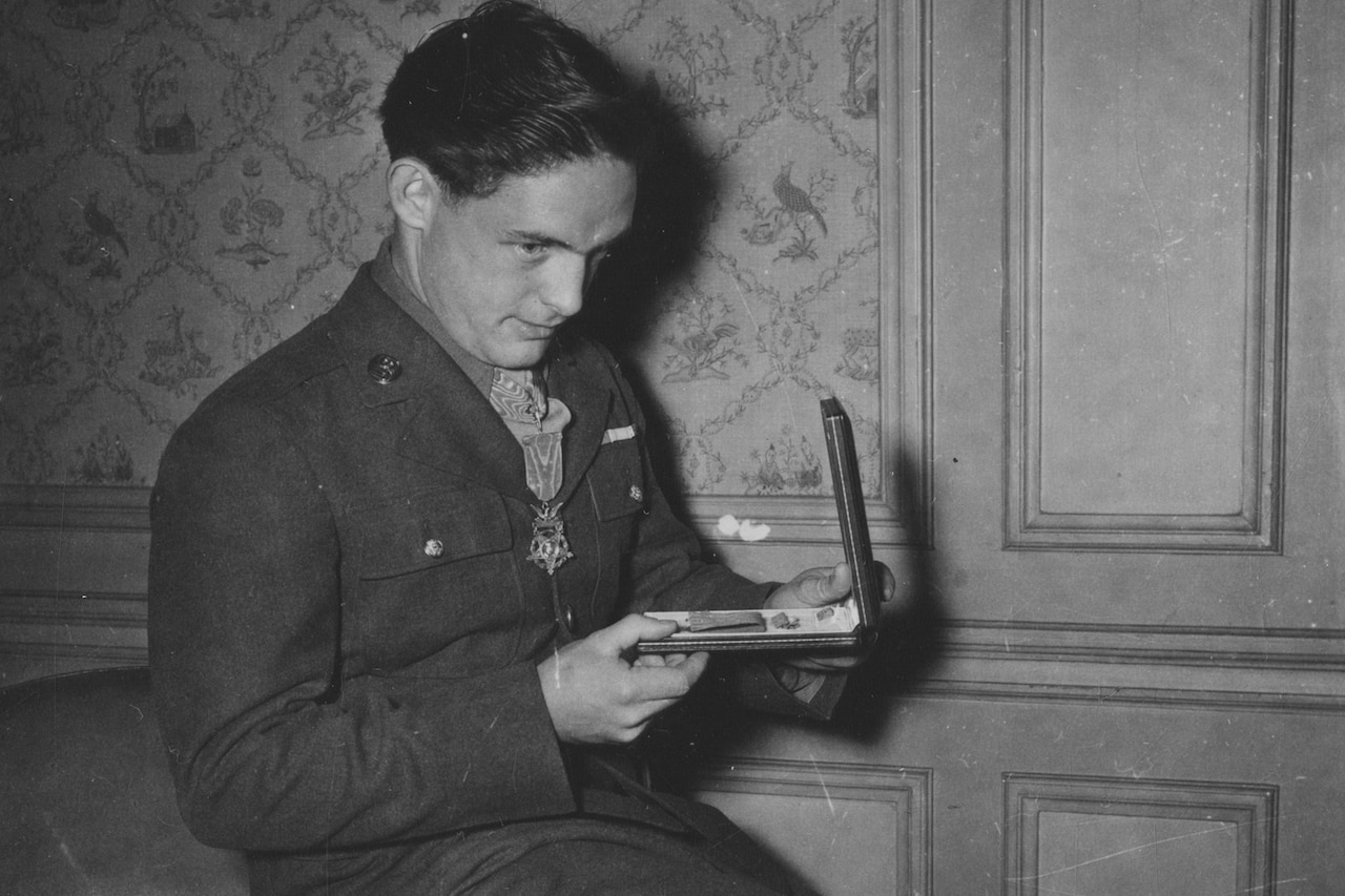 A person in a military uniform sits on the arm of a couch while looking down at a medal in a box.