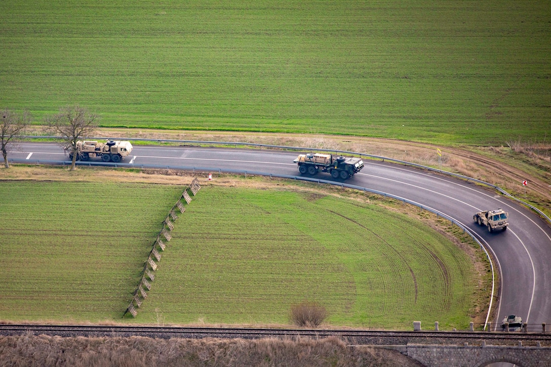 Four tactical vehicles turn a corner while traveling towards an opening under a bridge surrounded by fields as seen from above.