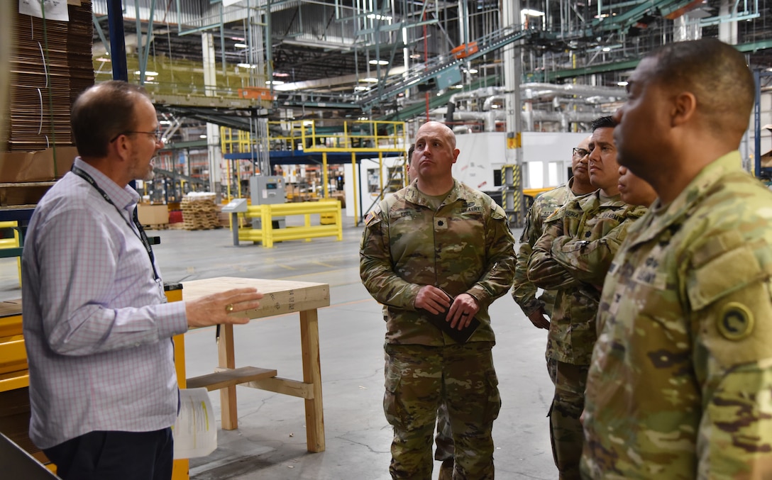 Photo is of a group of men, some in Army military uniform standing in a large warehouse.