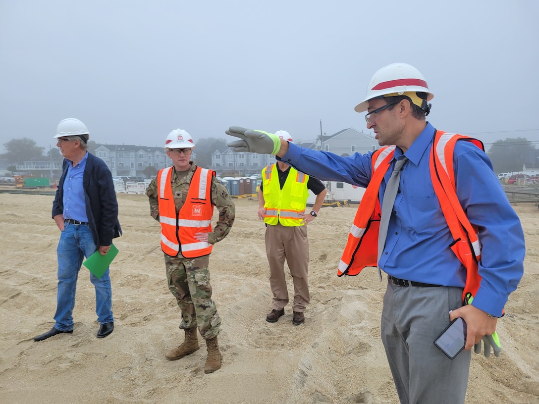 A man in a hardhat points at something in the distance while three other men wearing hardhats look onward. They are standing on a beach.