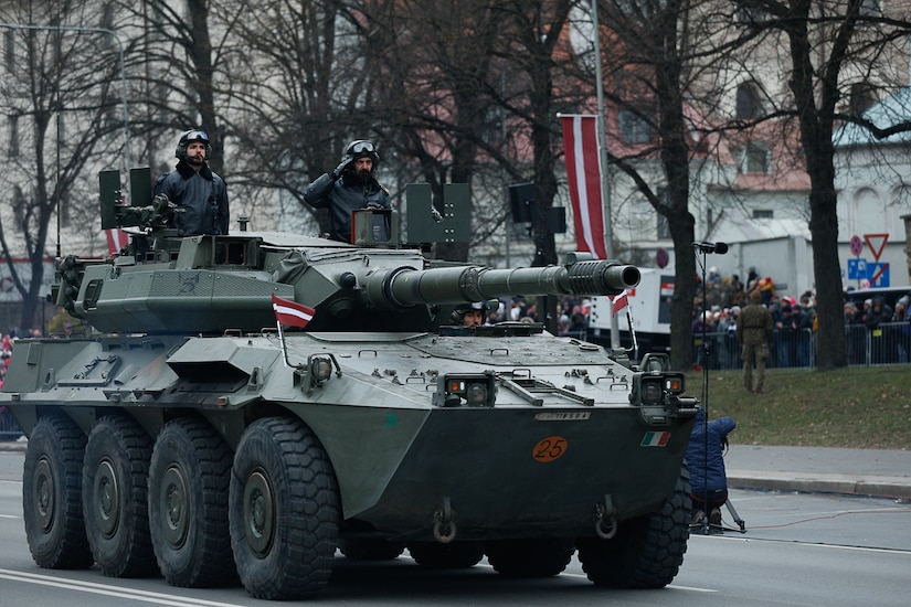 Foreign military members salute while riding aboard a military vehicle.