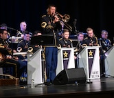 A soldier in a dark ceremonial uniform is playing a trombone in front of other band members who are seated behind music stands that have the words U.S. Army and Army Blues on them.