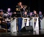 A soldier in a dark ceremonial uniform is playing a trombone in front of other band members who are seated behind music stands that have the words U.S. Army and Army Blues on them.