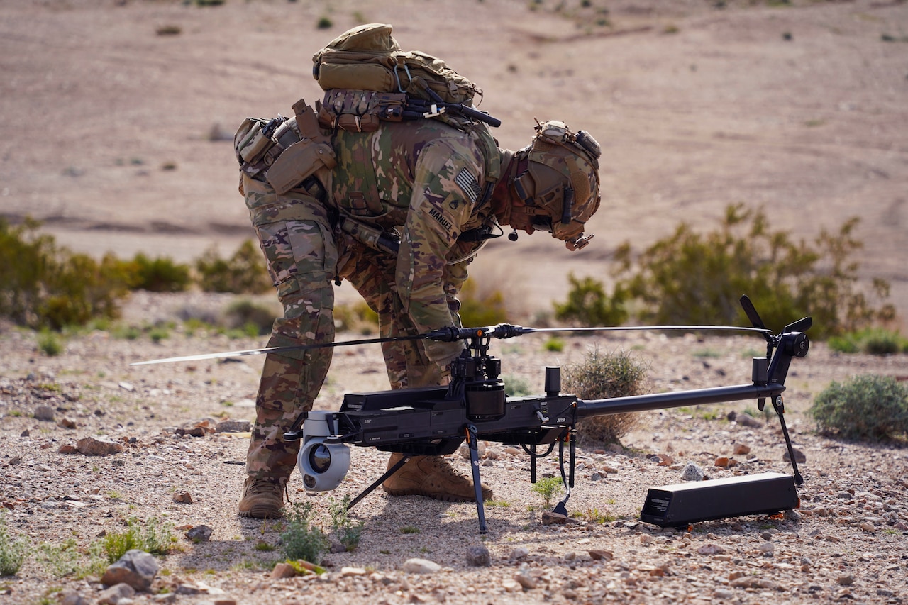 A soldier crouches in a desert area and prepares a human machine integration device with bushes scattered in the background.