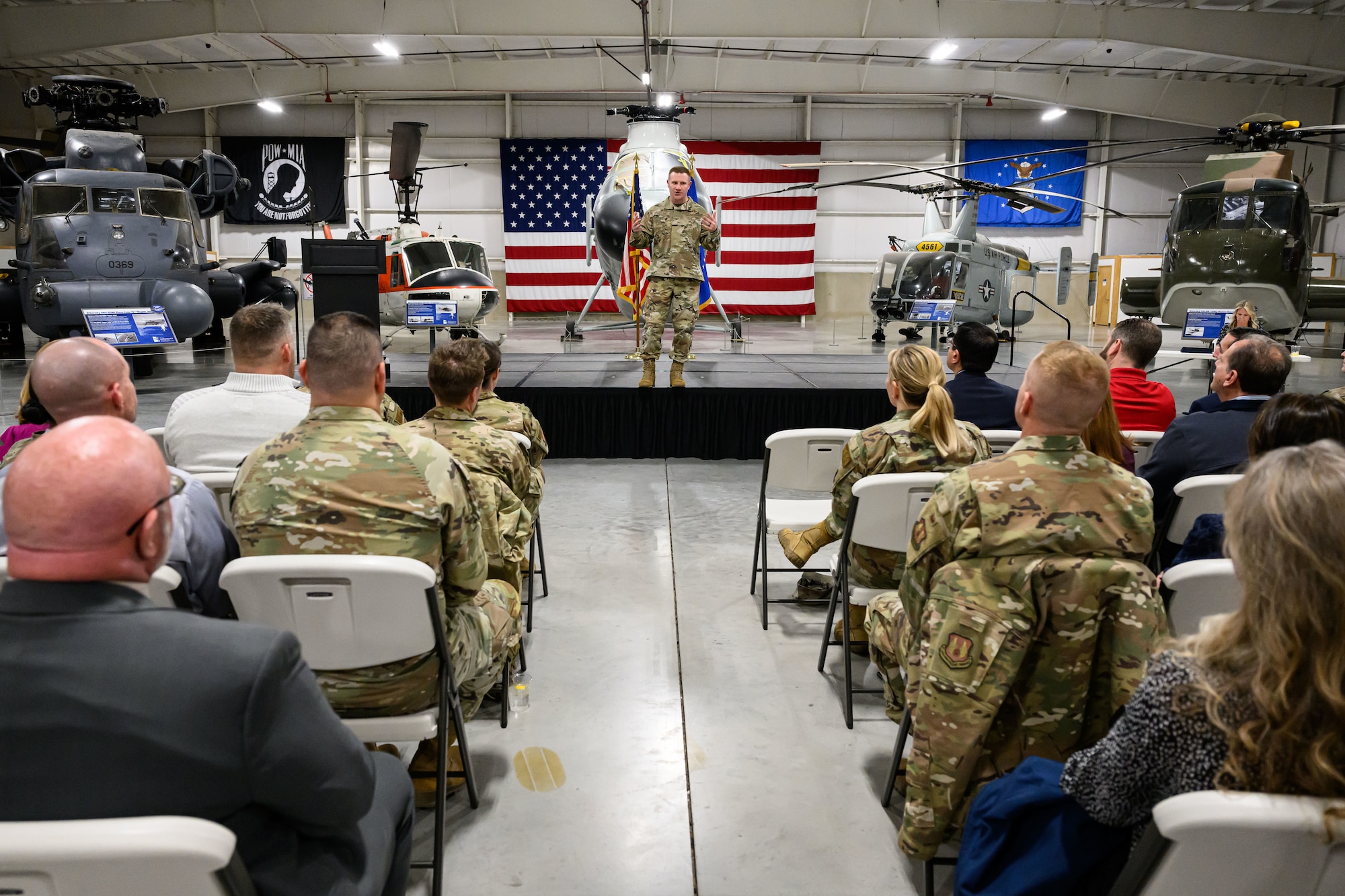 Col. Holland addresses a seated audience while atop a stage. Displays of aircraft in the museum are seen in the background.
