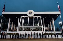 Army soldiers in dark ceremonial uniforms are- holding various musical instruments and are standing in a straight row directly in front of the Presidential Inaugural Parade reviewing stand, which is filled with people and has white trim and large windows. It is flanked by two US flags flying from white flagpoles, and at the top of the reviewing stand is the dark blue official seal of the U.S. President.