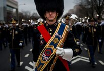 Army Soldier dressed in a band drum major uniform (large dark furry hat with strap, blue sash with yellow border across chest with colorful military ribbons and insignias, red sash underneath going across chest on opposite shoulder, carrying large wooden and silver-tipped mace in front in one white-gloved hand) is marching in front of other Army Soldiers in dark uniforms, some carrying musical instruments and performing.