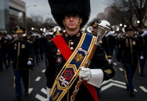 Army Soldier dressed in a band drum major uniform (large dark furry hat with strap, blue sash with yellow border across chest with colorful military ribbons and insignias, red sash underneath going across chest on opposite shoulder, carrying large wooden and silver-tipped mace in front in one white-gloved hand) is marching in front of other Army Soldiers in dark uniforms, some carrying musical instruments and performing.