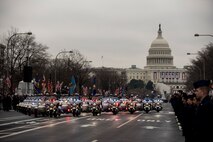 Dozens of police motorcycles and other vehicles are driving down a street away from the U.S. Capitol building. Their red and blue lights are flashing. There are people lining the street watching.