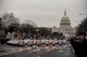 Dozens of police motorcycles and other vehicles are driving down a street away from the U.S. Capitol building. Their red and blue lights are flashing. There are people lining the street watching.