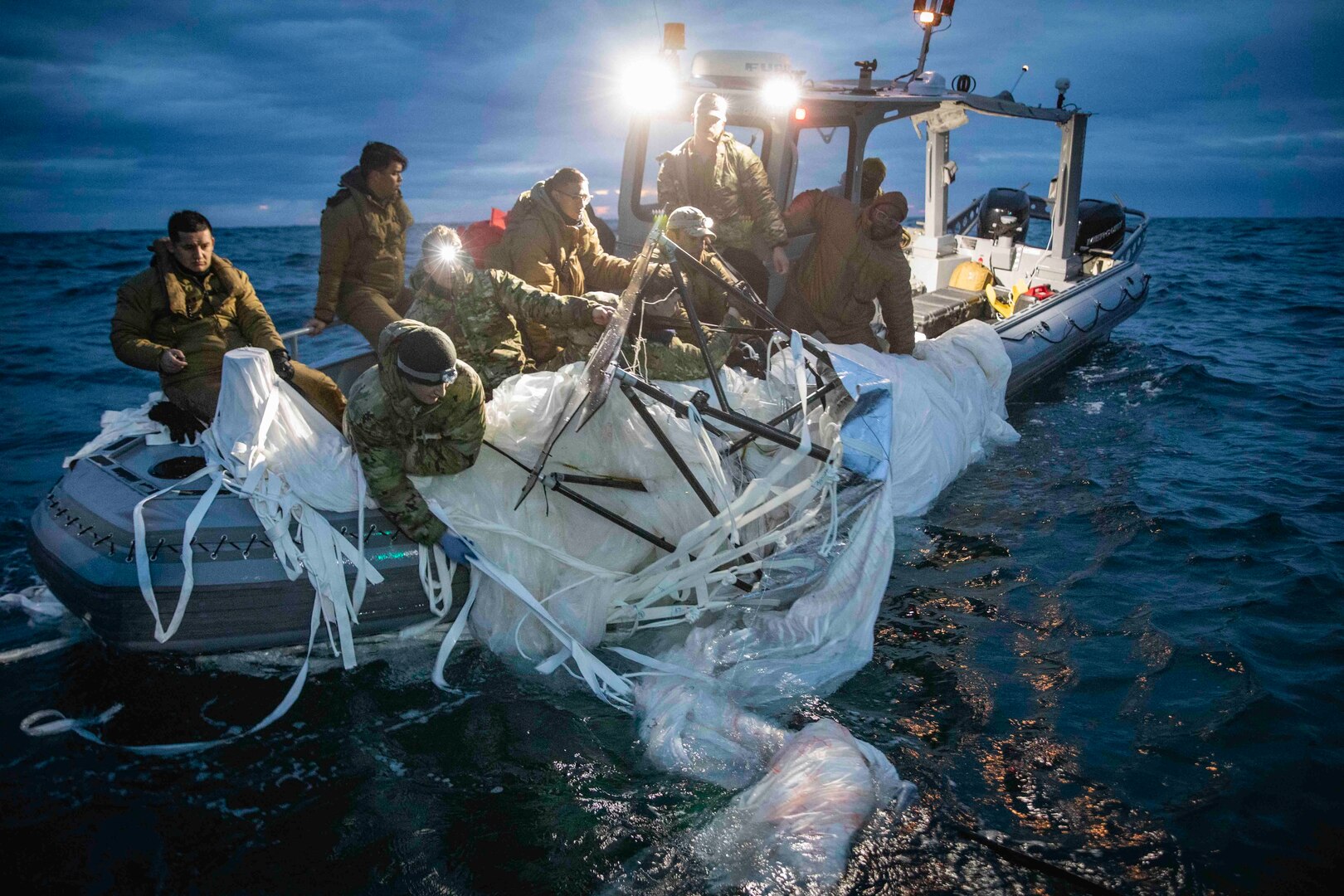 About eight service members in camouflage are in a small boat pulling in a deflated plastic balloon at night.