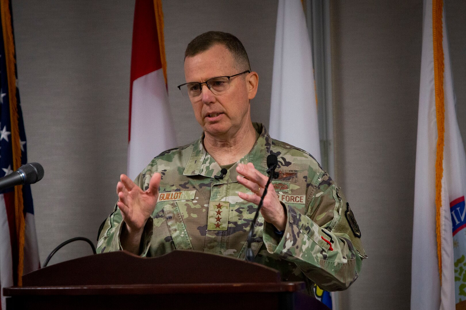 A man in camouflage stands at a lectern with his hands at chest level.