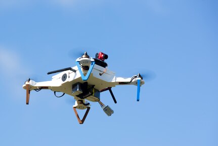 A square, plastic aircraft with a spinning rotor on each corner flies in the air with a clear blue sky behind it.