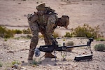 A soldier crouches in a desert area and prepares a human machine integration device with bushes scattered in the background.