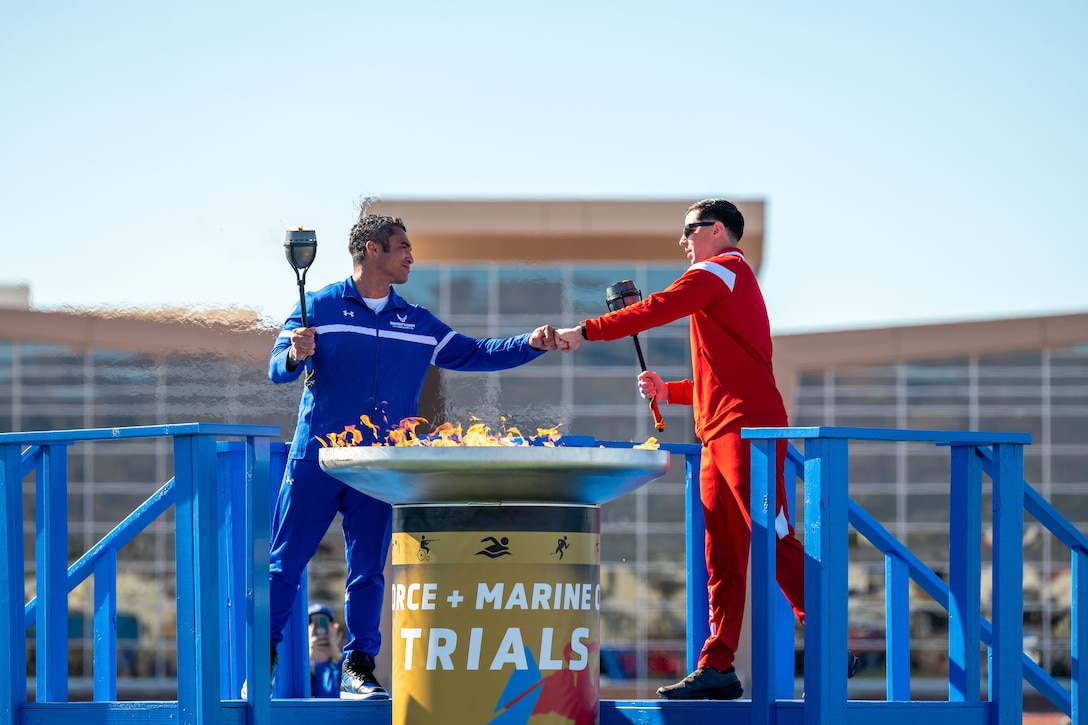 A retired airman and a Marine dressed in athletic gear and holding small torches bump fists while standing on a blue wooden platform. A large torch burns in front of them.