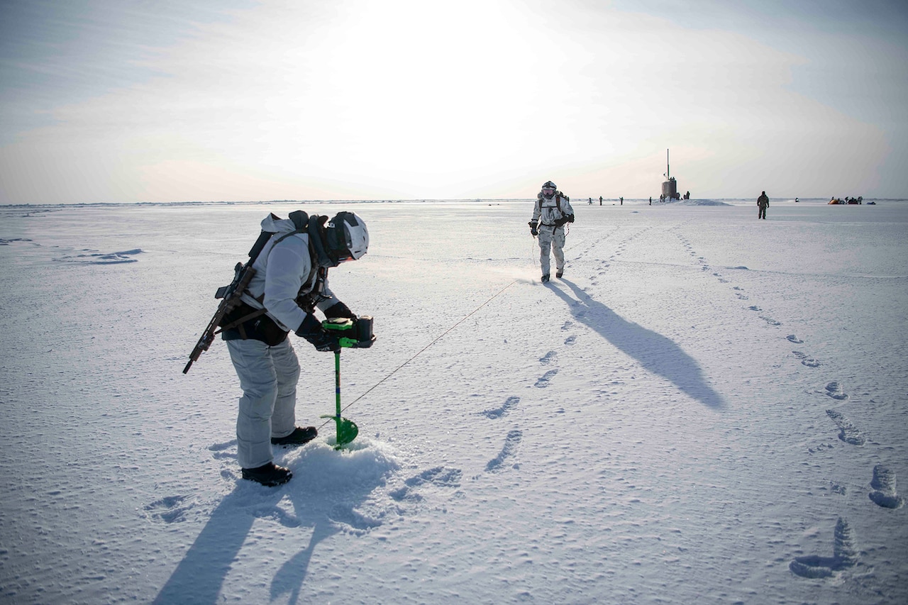 A sailor uses an ice auger during daylight to cut into snowy ground as another sailor approaches. A submarine can be seen in the distance.